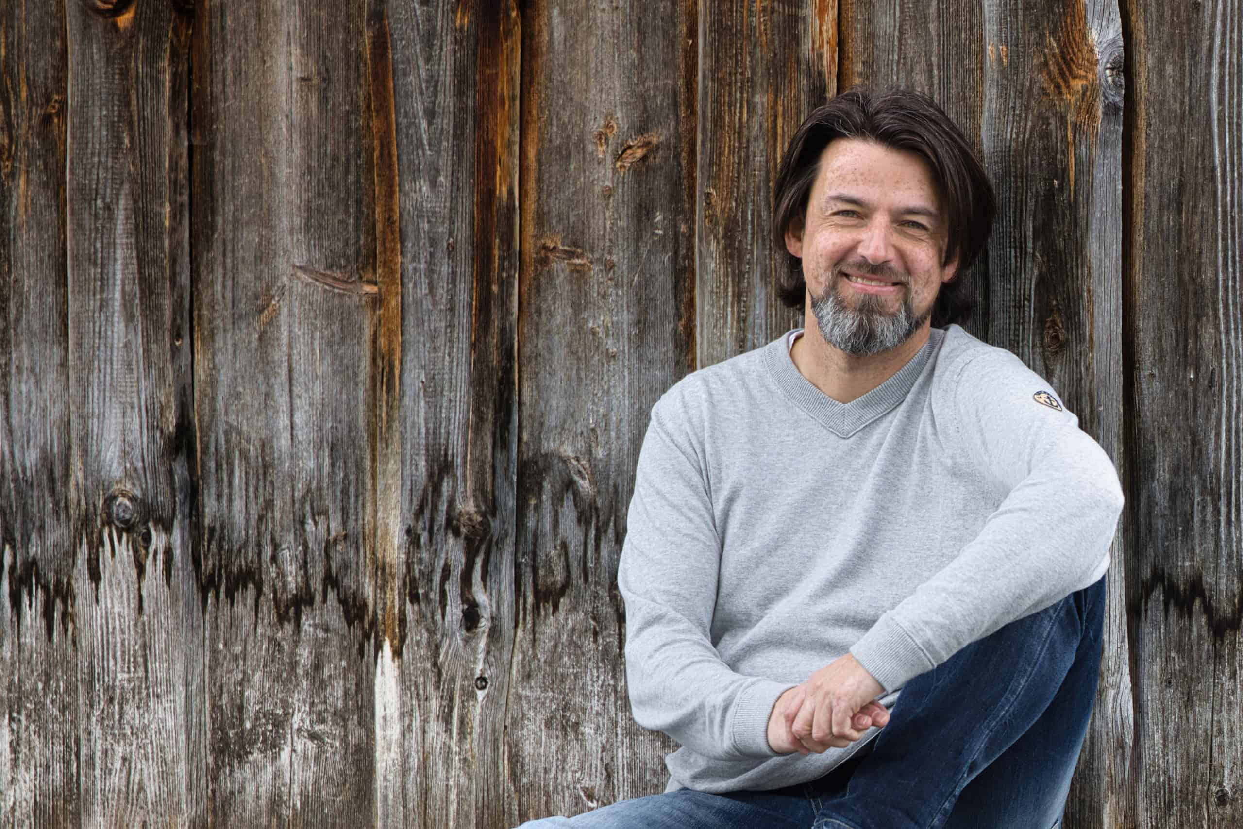 Heilkreis und Energiearbeit mit Oliver Hofmann Mann sitzt entspannt vor einer Holzwand, symbolisiert Heilkreis und energetische Arbeit, vermittelt Ruhe und innere Balance.